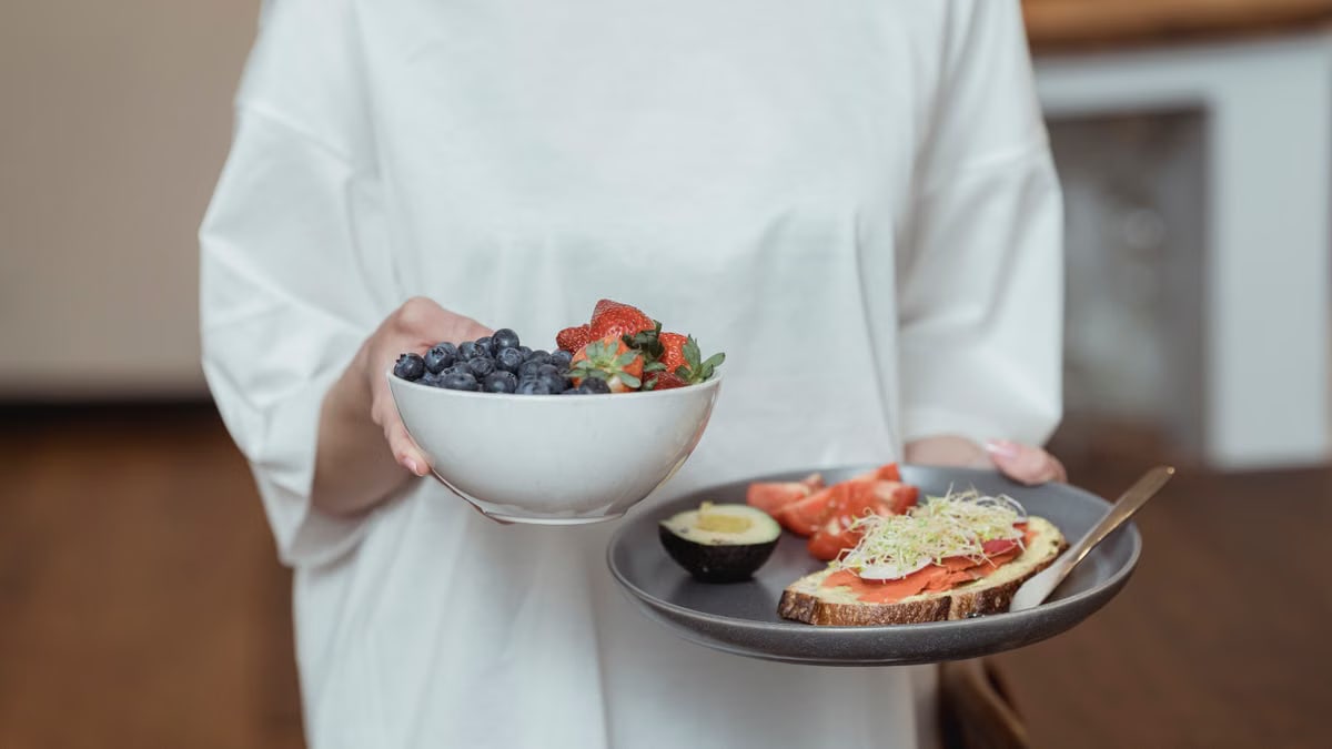 Person holding a bowl of fresh berries and a plate with avocado toast, displaying a healthy breakfast spread.
